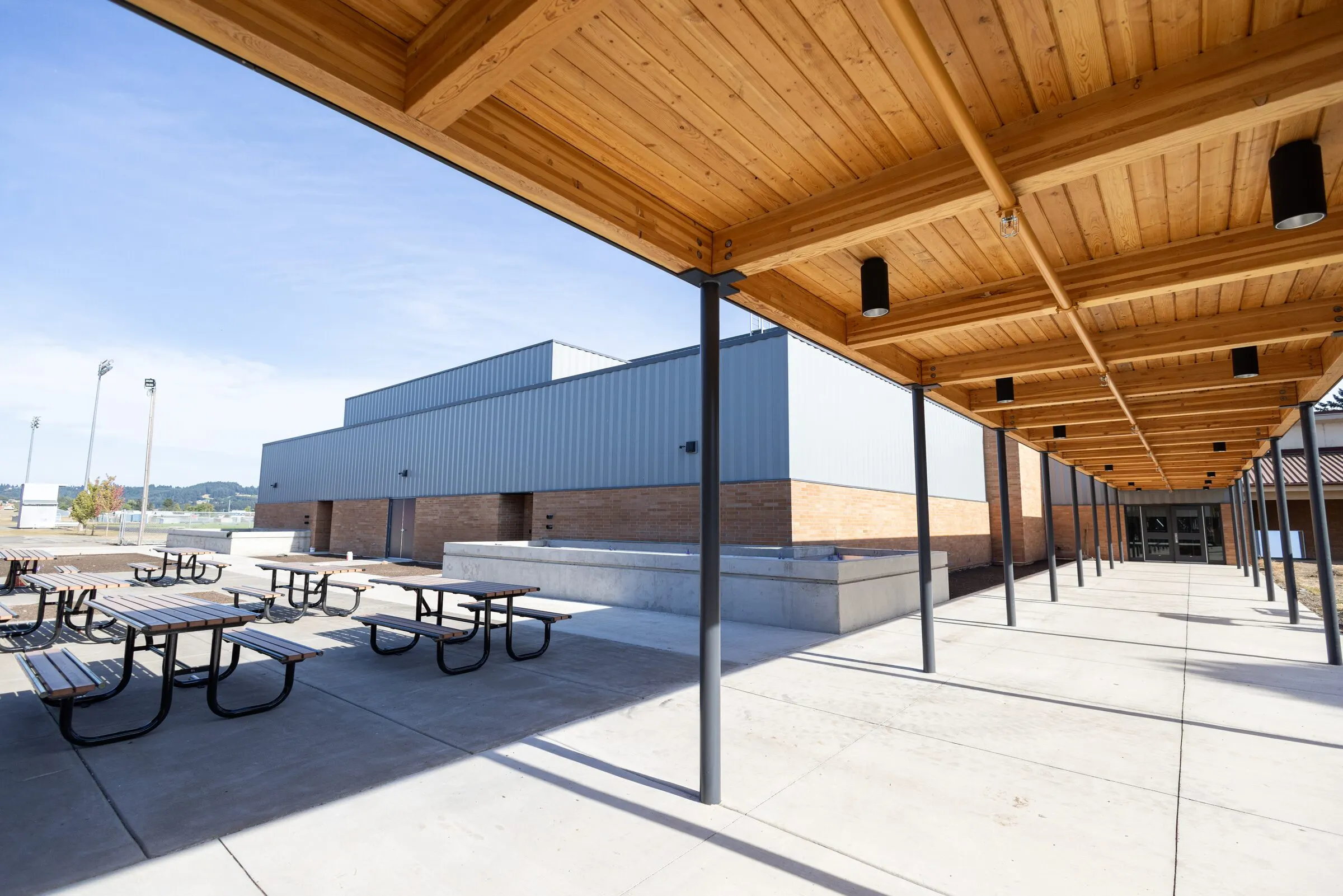 Outside the new Amity High School on a clear day — a long covered walkway with a mass-timber ceiling, brick columns, picnic benches, and the new athletic wing across the courtyard.